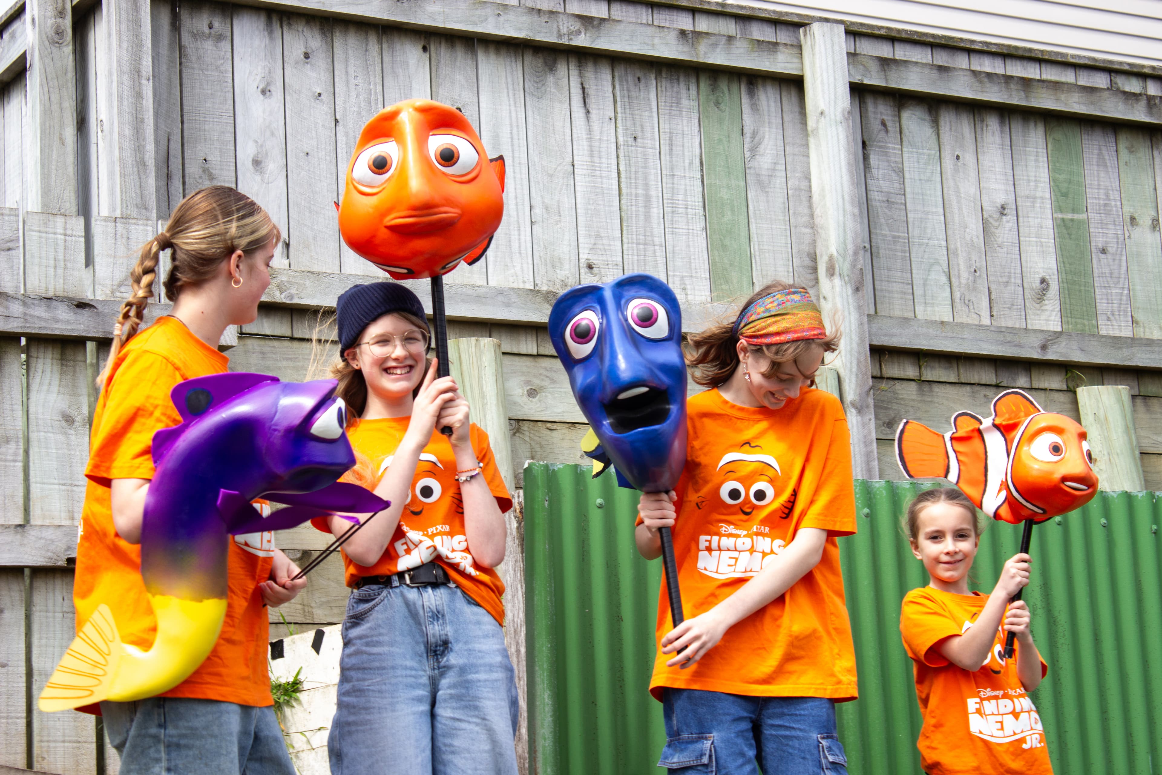 four smiling children wearing orange tshirts which have a picture of Nemo and the words "Finding Nemo" are holding large rod puppets from the animated film "Finding Nemo". There is Gurgle looking at the other puppets from the side, Martin looking worried, Dory looking a bit confused but happy with mouth open as if mid song and Nemo looking off to the side. Like in the film the characters are bright, with exagerated facial features and large eyes