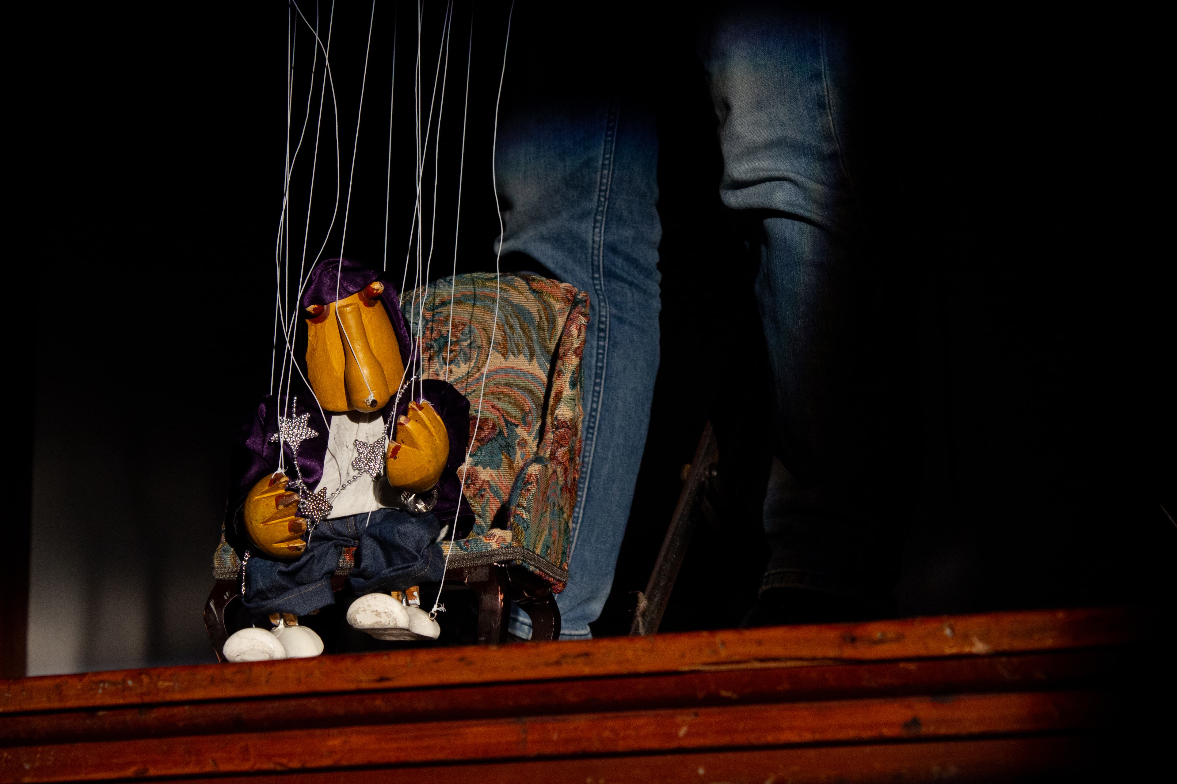 a wooden marionette wearing a purple velvet hoodie, white tee-shirt silver star necklace, blue jeans and white shoes and sits in a chair. He has a stylised cylindrical face with a long nose and two red beady wooden eyes protruding just below the hoodie. The puppet's head is slightly tilted one hand is gesturing towards the audience. The strings and puppeteers legs are visible.