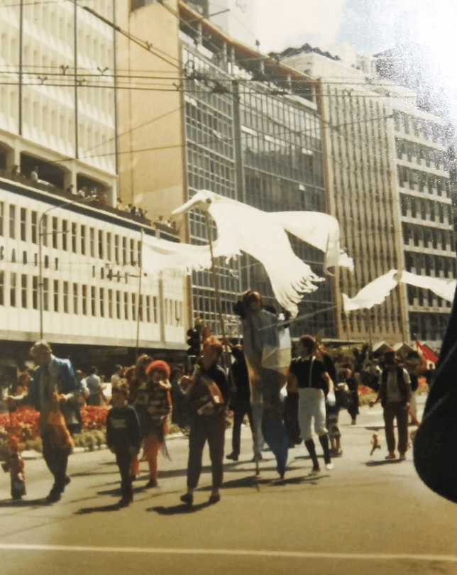 A large white bird parade bird puppet is held on rods by puppeteers at a parade down 1980s Lampton Quay Wellington