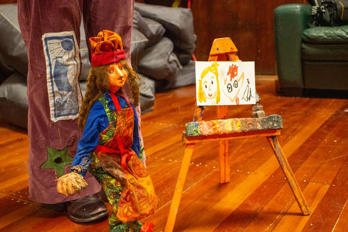 A marionette dressed as a portrait artist, with curly slighly matted brown hair and paint covered hands. She is wearing an orange and red paint spattered apron and a matching beret, sitting next to an easel displaying her work. On the orange easle which is covered with paint is a small canvas with her childlike signed painting of a smiling face with long blod heair and a person with short red hair."