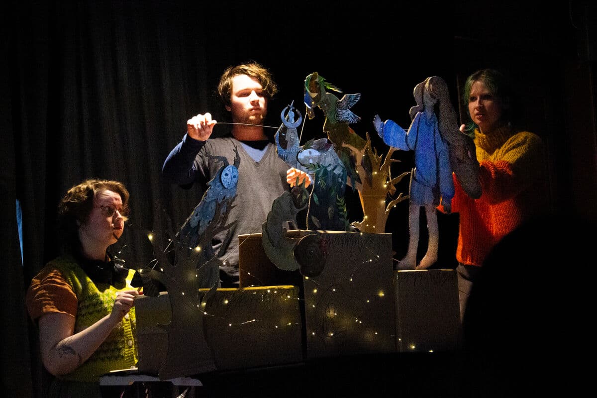 In a dark room there are 3 puppeteers. A beam of light illuminates the middle puppeteer who is looking intently at the flat rod puppet of a fantastical creature in front of him. It looks like a wood sprite emerging from the tree. To his left a puppeteer with a worried expression looks towards the 2 dimensional rod puppet of a girl she is operating. On the other side, kneeling a puppeteer looks toward the girl puppet which her puppet, another fantastical creature that has a moon face and antlers and tree branch arms is looking towards. The puppets are bathed in blue light and are performing on black boxes wreathed in fairy lights.