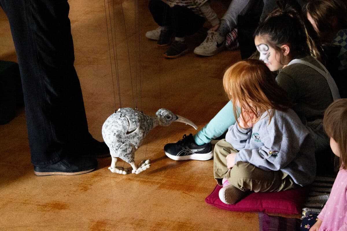 A wooden kiwi marionette with one foot raised is approaching two children who are sitting on the floor and smiling. The puppet's feet are beautifully carved you can see each tonail on the foot and it has a long curved beak, grooves where wings will fold up from are visible. The kiwi is painted in browns and white with a light yellow beak and feet.