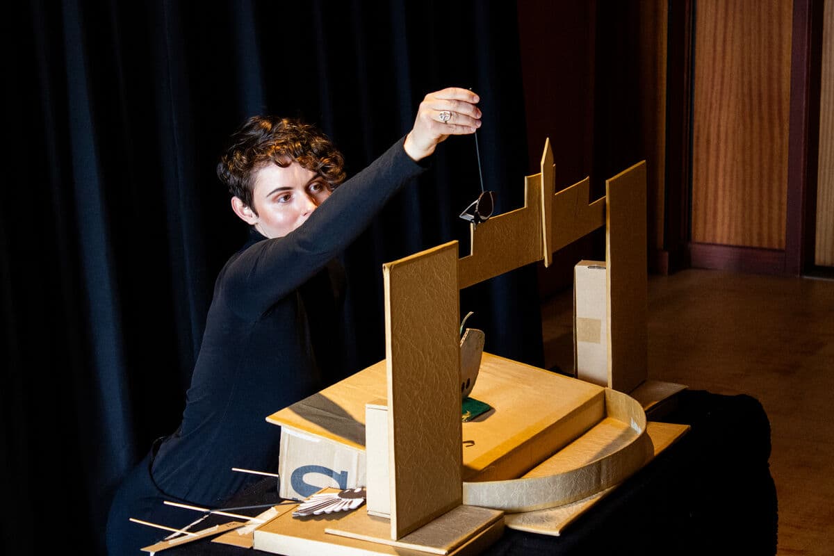 puppeteer with brown curly hair and dressed in black is performing a puppet show taking place in a mini table top theatre ,made out of plain brown cardboard. The puppeteers mouth is obscured by her raised arm and her eyes are focused on the puppet she is operating. The puppet centre stage is a rock. The bird puppet lying down off stage is visible.