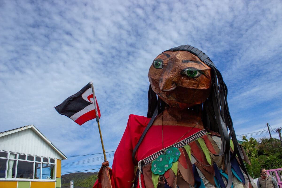 Standing in front of a bright blue sky streaked with white clouds is large parade style puppet of a Māori woman wearing a red top and a korowai over one shoulder. She appears enourmous dwarfing the house in the background. She carrys a tino rangatira flag. The puppet is made from willow with coloured tissue paper over the frame. The shape of willow can be seen underneath the tissue papper. Her hair is made from black tissue paper hanging in long ropes down below her shoulders.