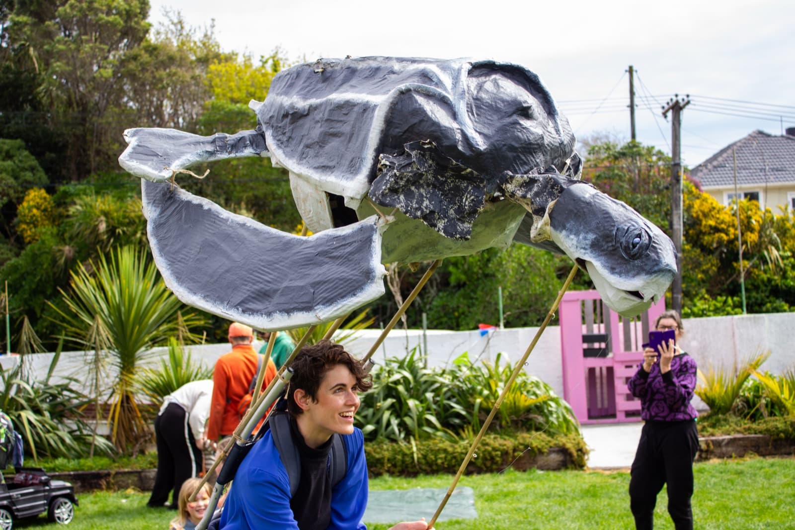 A puppeteer wearing a backpack style turtle puppet operated with bamboo rods. Puppet is made from papier mache painted grey and has rubbish stuck to it's underside.