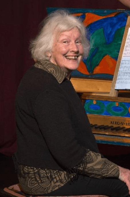 A pakeha woman in her 60s with white hair sitting at a brightly coloured harpsichord and smiling.ivals