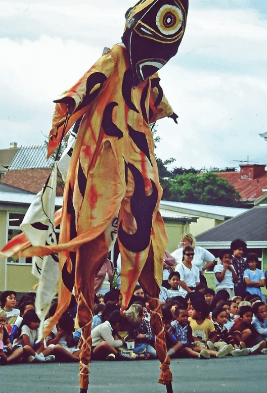Stilt performers in a flame coloured costume with a stylised double sided red head mask with large round white and yellow eyes