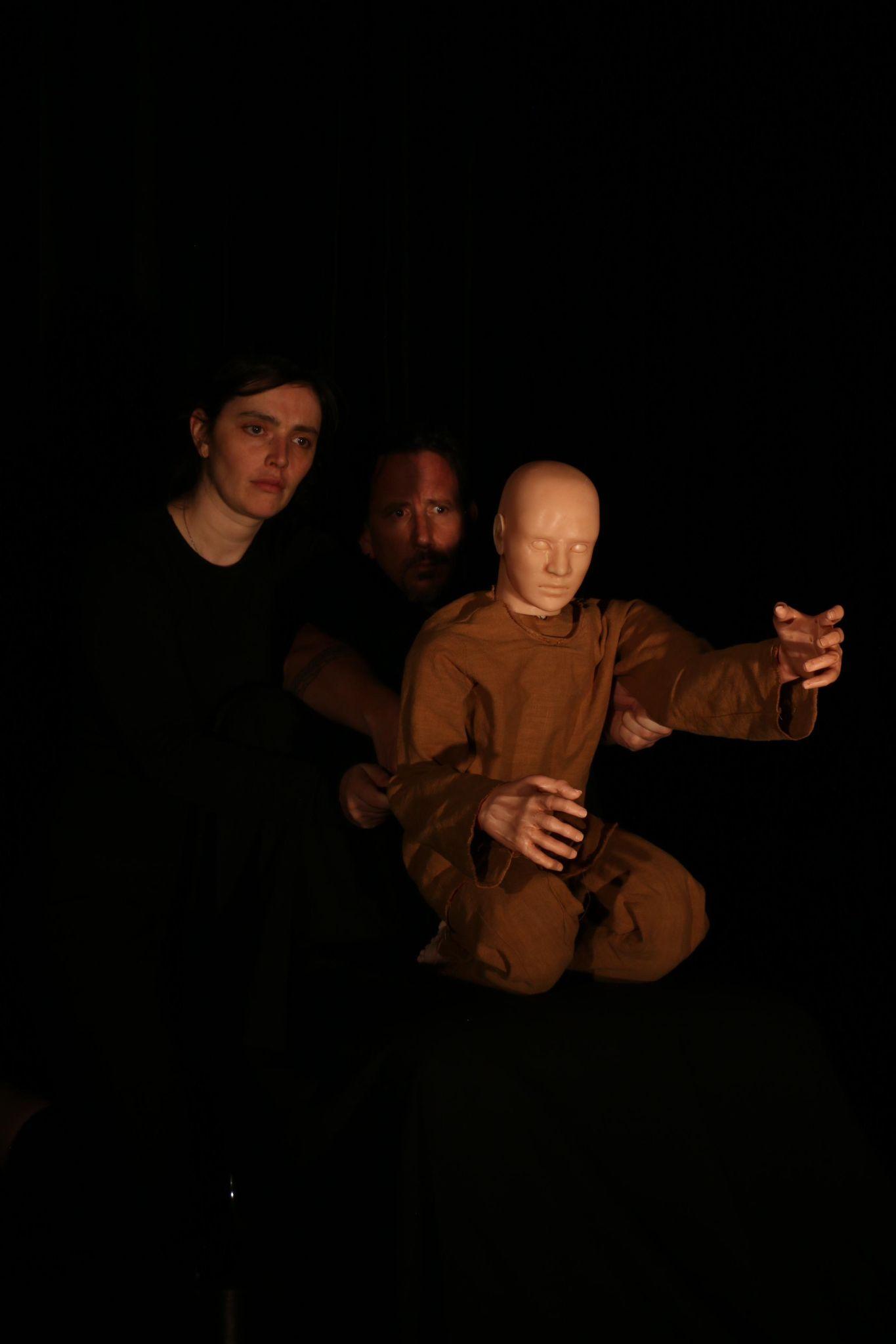 Two performers dressed in black kneel in darkness, operating a life-size child puppet in ochre-coloured clothing as it reaches one arm forward into the light.