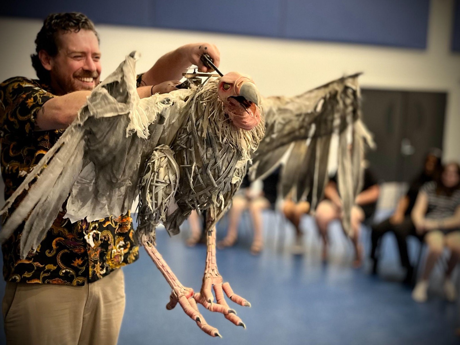 Jon Coddington smiling while operating a large vulture puppet with tattered wings, in front of a seated workshop audience