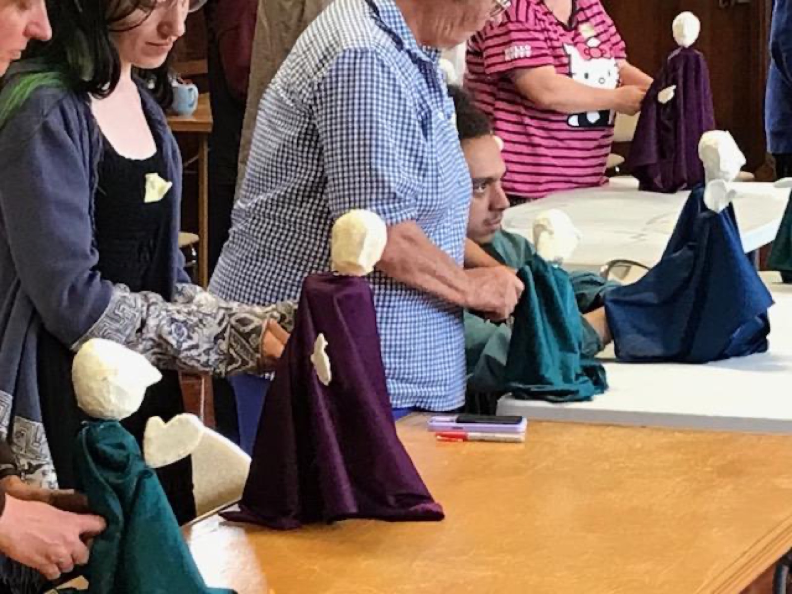 Workshop participants standing at a table, each holding a simple puppet with a white foam head and dark fabric cape they have just made