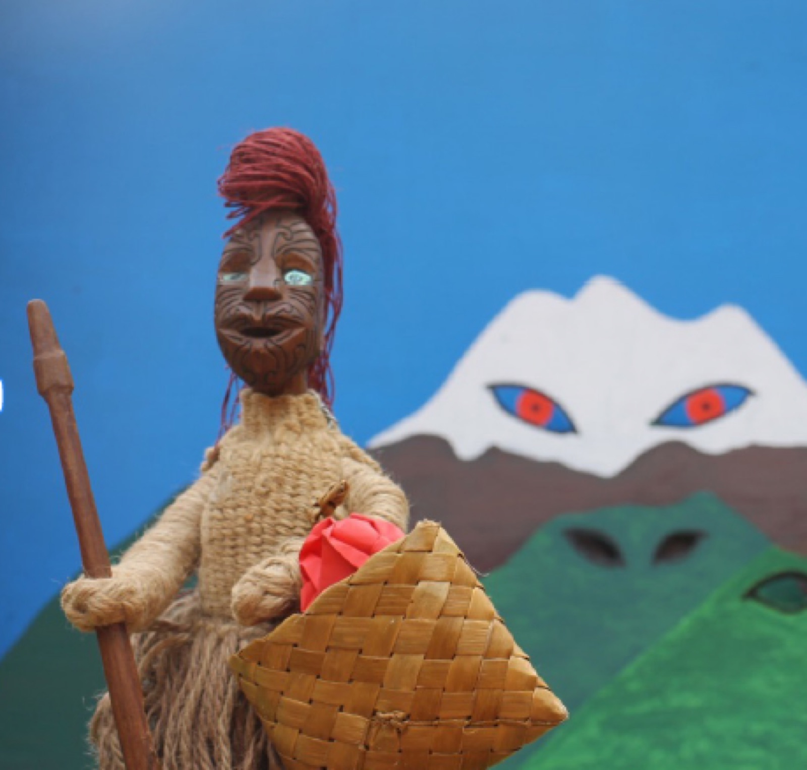 A carved kare-tao puppet with a moko, red hair and a woven kete, held up in front of a painted backdrop of a snow-capped maunga with red eyes