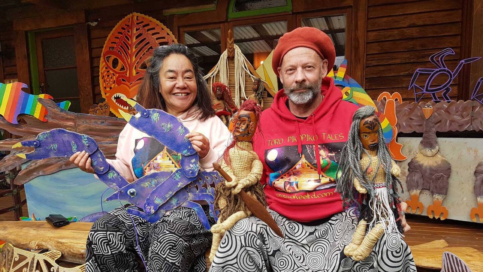 Smiing puppeteers, Whaitaima Te Whare and Jeffrey Addison sitting side by side holding carved and painted puppets, including a blue ‘flatsie’ taniwha, in front of a carved and brightly painted wall