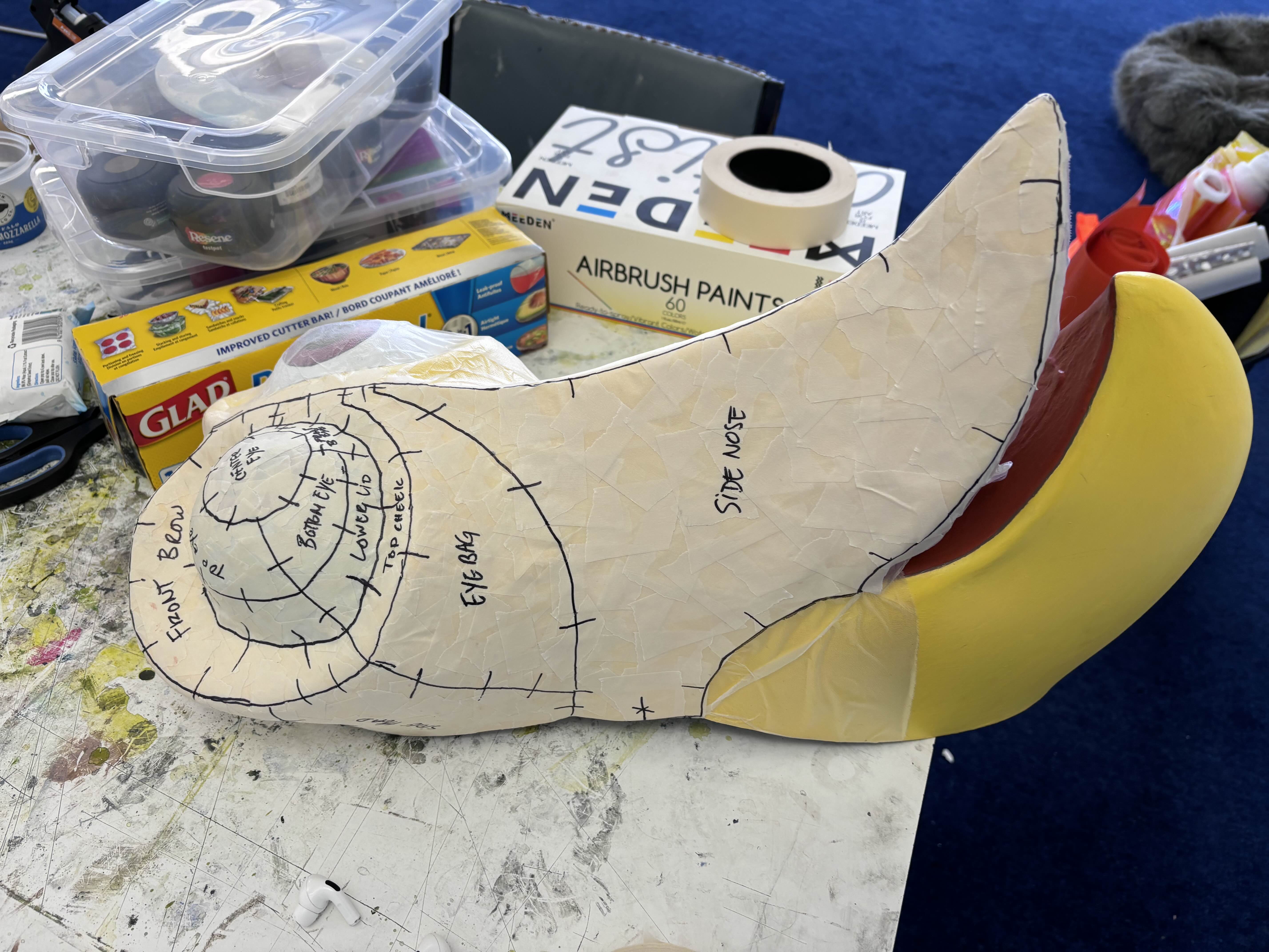 A pale foam head sculpt marked up with pattern lines and labels such as 'front brow', 'eye bag' and 'side nose', sitting on a workbench surrounded by craft supplies.