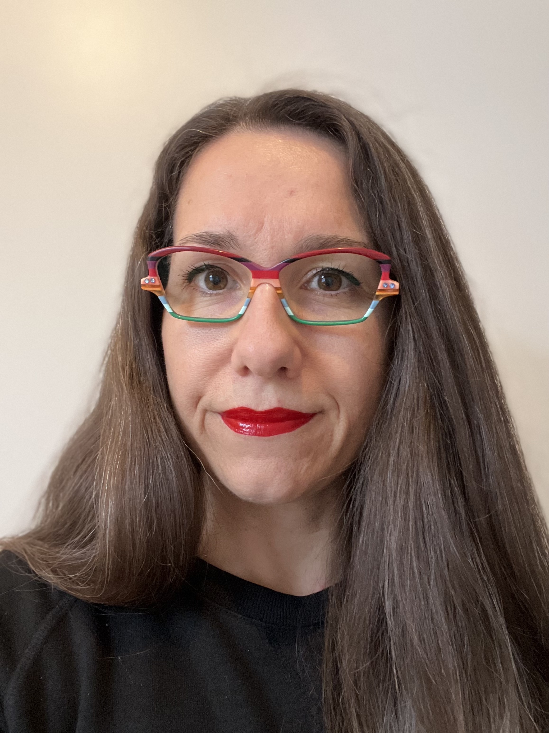 Portrait of Sarah Bell, a woman with long brown hair wearing colourful patterned glasses and red lipstick.