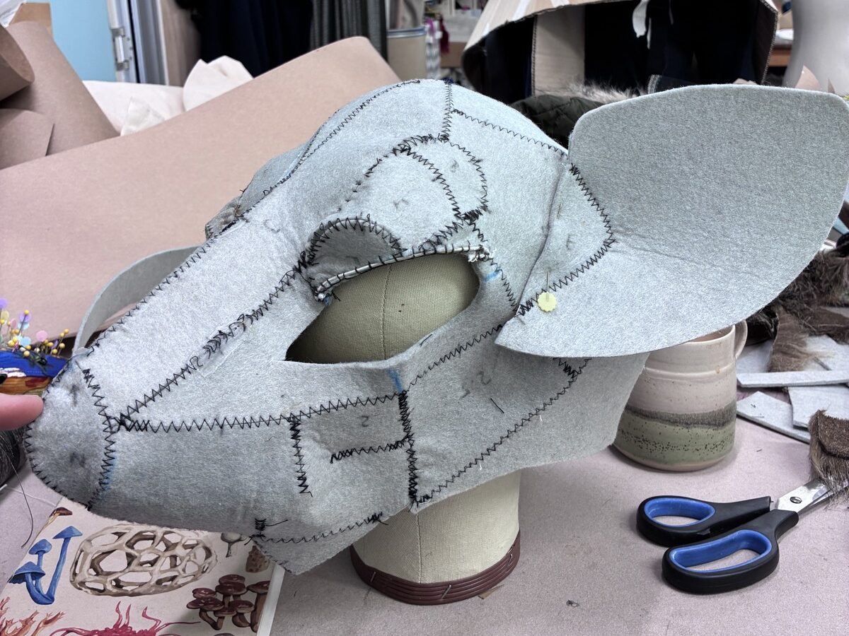 A grey felt rat head, machine-stitched together from flat pattern pieces, fitted over a dressmaker's head form in a busy workroom.