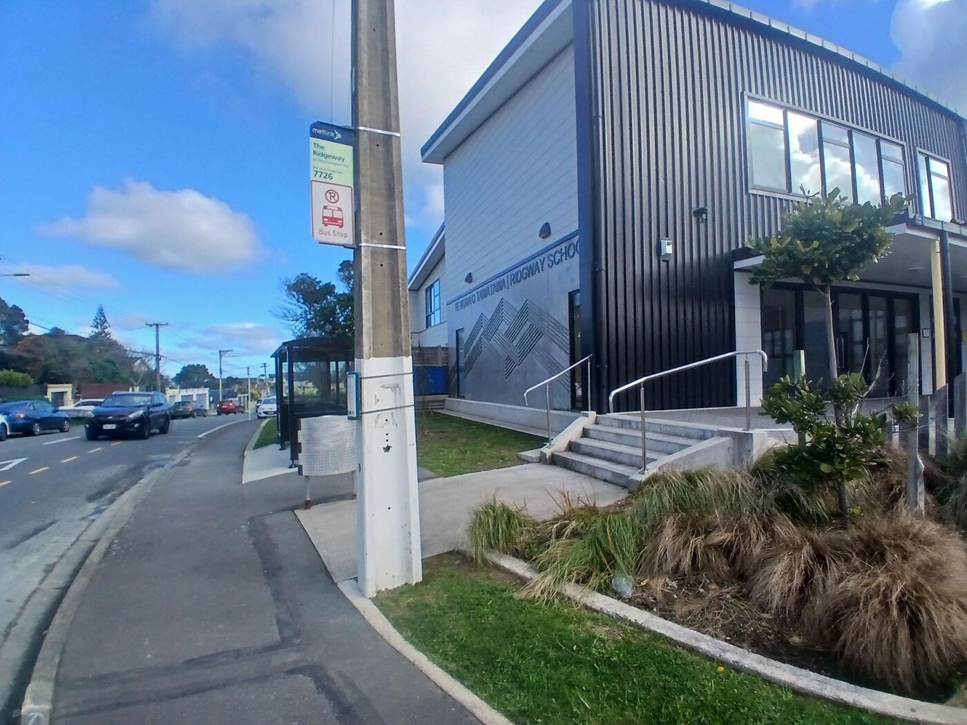A Metlink bus stop pole numbered 7726 on the footpath on Mornington Road at Ridgway School, with a small glass-sided shelter behind it. Concrete steps with a metal handrail lead up from the footpath.