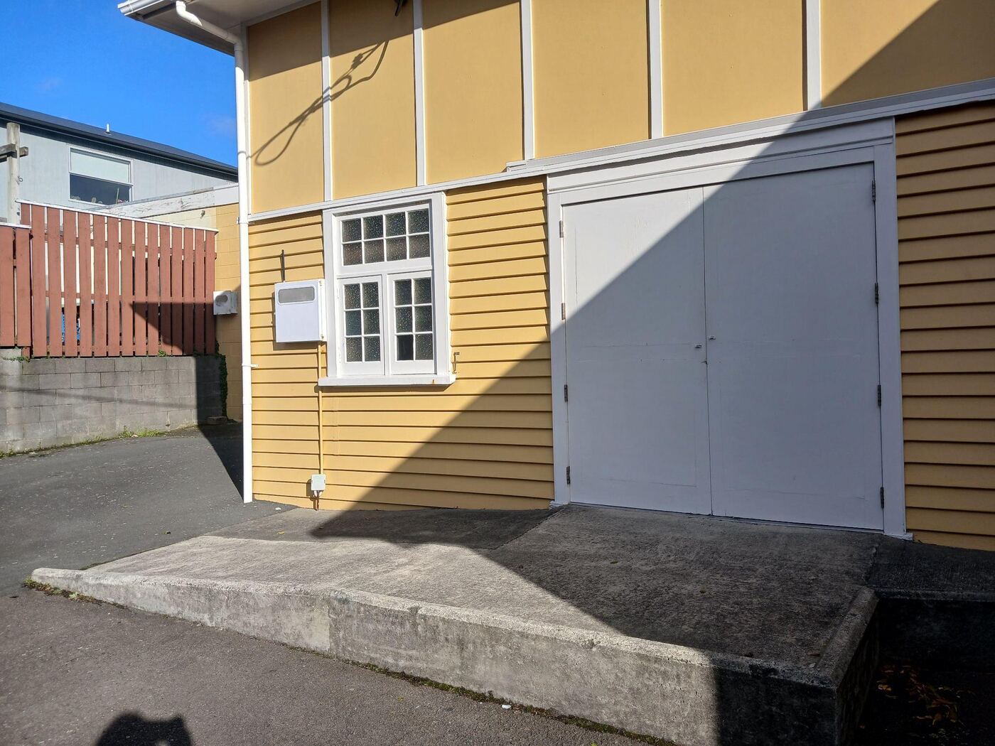 The same doors seen from outside: a pair of light-coloured double doors in the hall’s yellow weatherboard wall, opening onto a level concrete ramp with garden shrubs along its edge.