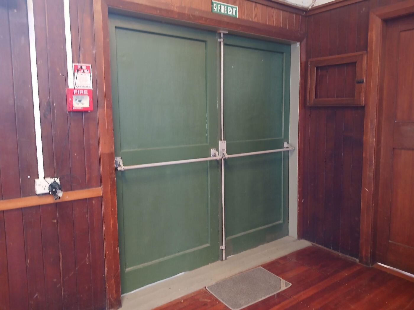 The accessible entrance seen from inside the hall: a pair of green double doors with a horizontal push bar, set into the dark wood-panelled wall near the stage, with a doormat on the floorboards in front of them.