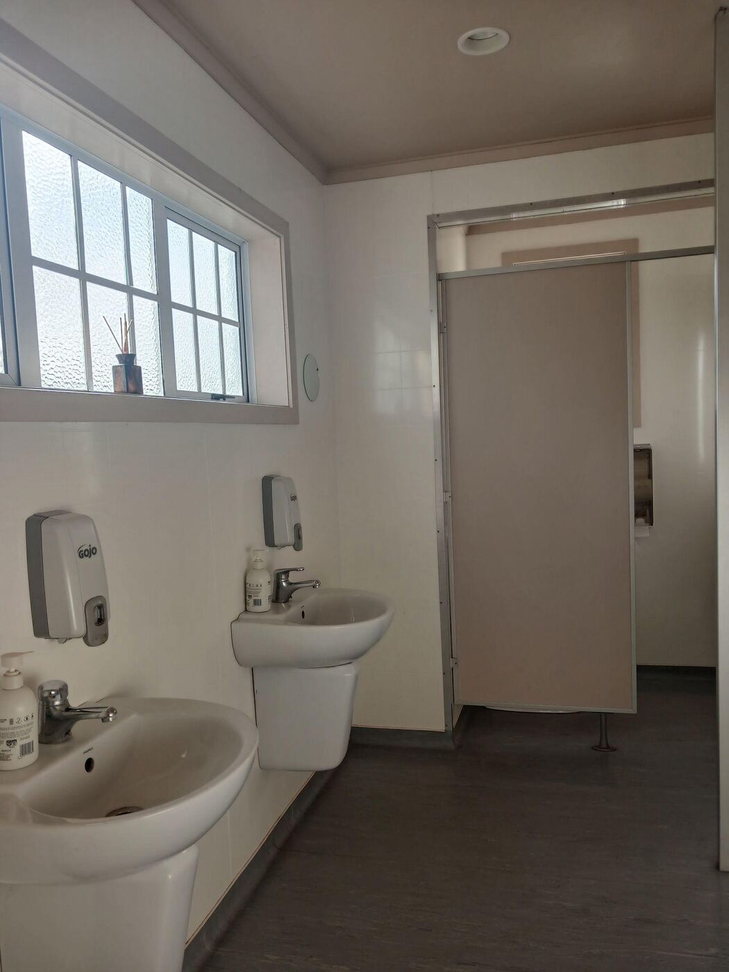 Inside one of the hall toilets: two wall-mounted sinks under a frosted window, soap dispensers and a towel rail. A partition wall with a sliding door leads through to a cubicle, and the floor is dark wood.