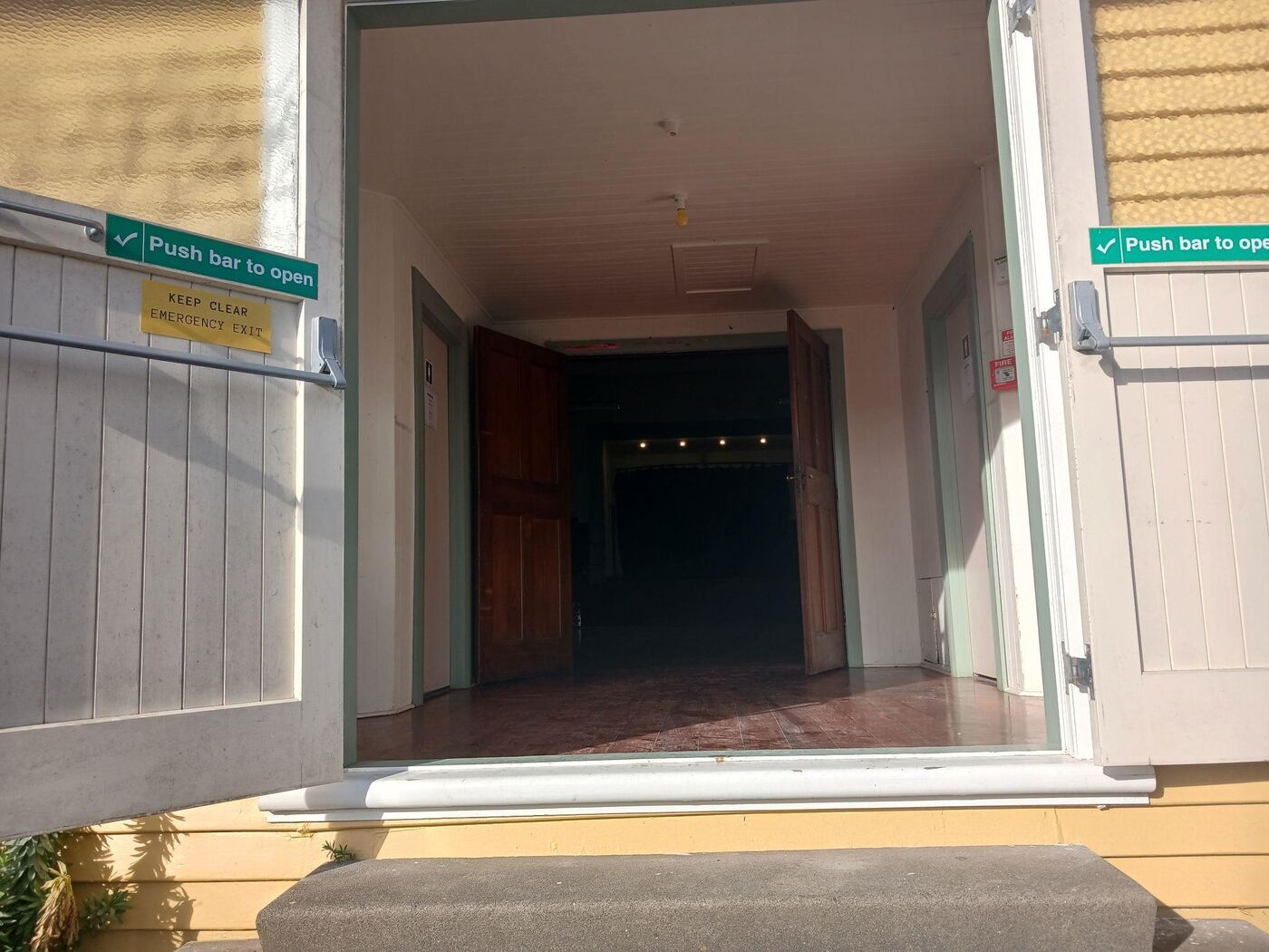 Looking through the open double doors at the top of the entrance steps into a dark hallway, with doors on either side and a fire exit sign overhead. There is a raised threshold to step over just inside the doorway.