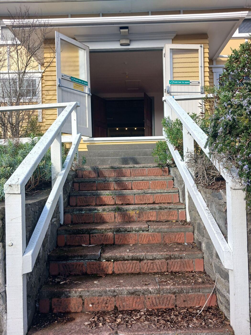 A flight of red brick steps with white-painted wooden handrails on both sides, leading up a garden path to open double doors into the hall. Low shrubs and a stone retaining wall line the steps on either side.