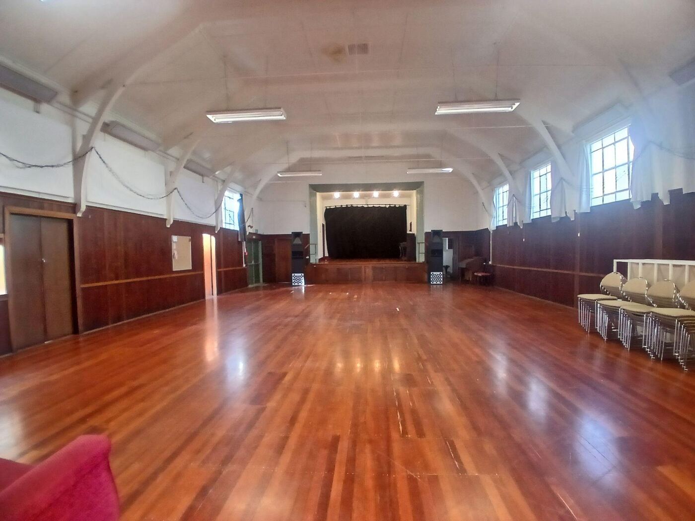 Inside the hall: a long room with a curved white ceiling, dark wood-panelled walls and polished wooden floorboards. A raised stage with a black curtain is at the far end, with tall windows down one side and stacked chairs along the other.
