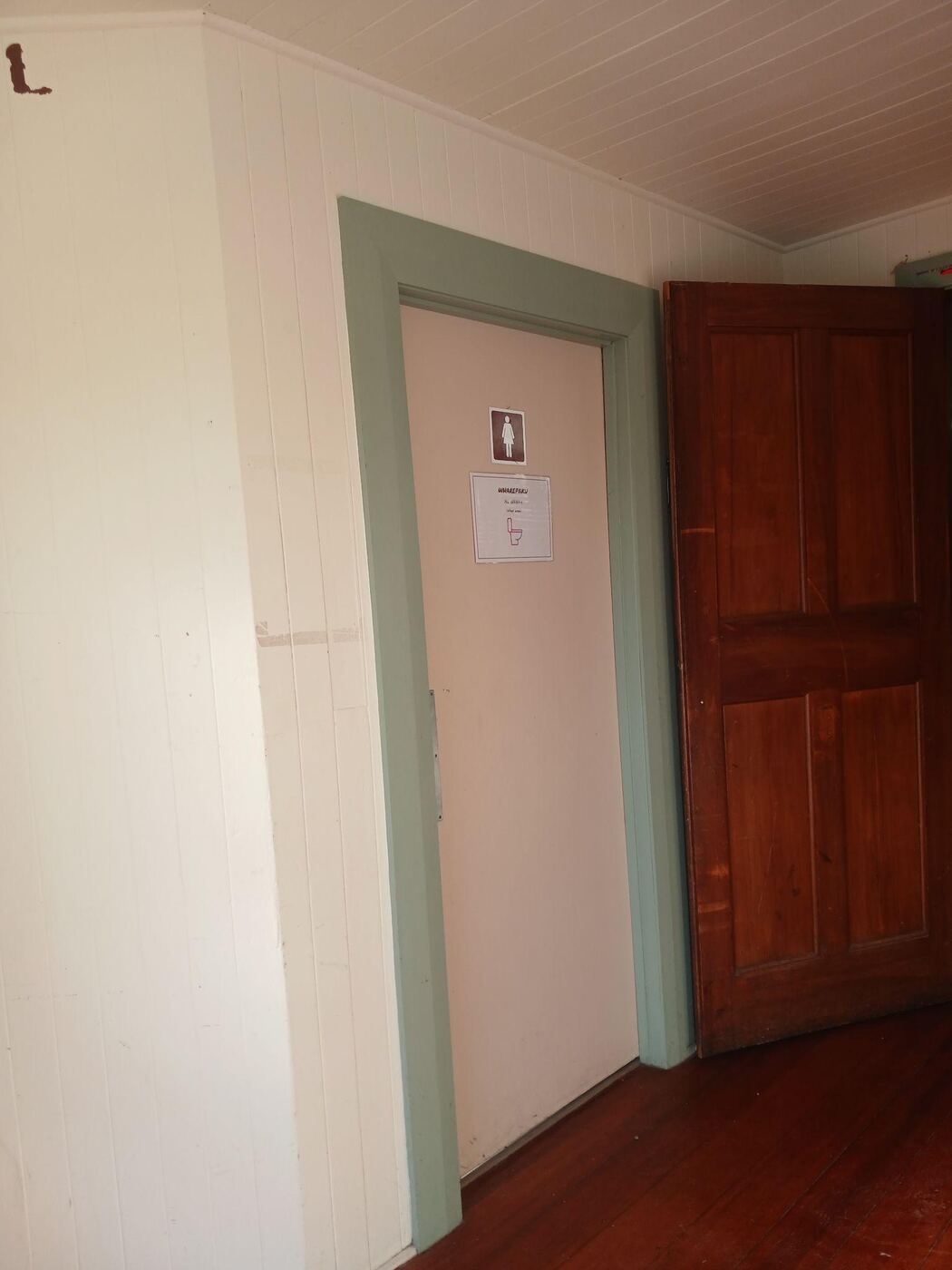 A toilet door off the foyer, marked Wharepaku with a female symbol, set into a pale green-panelled wall beside a large open wooden door. The floor is dark polished wood.