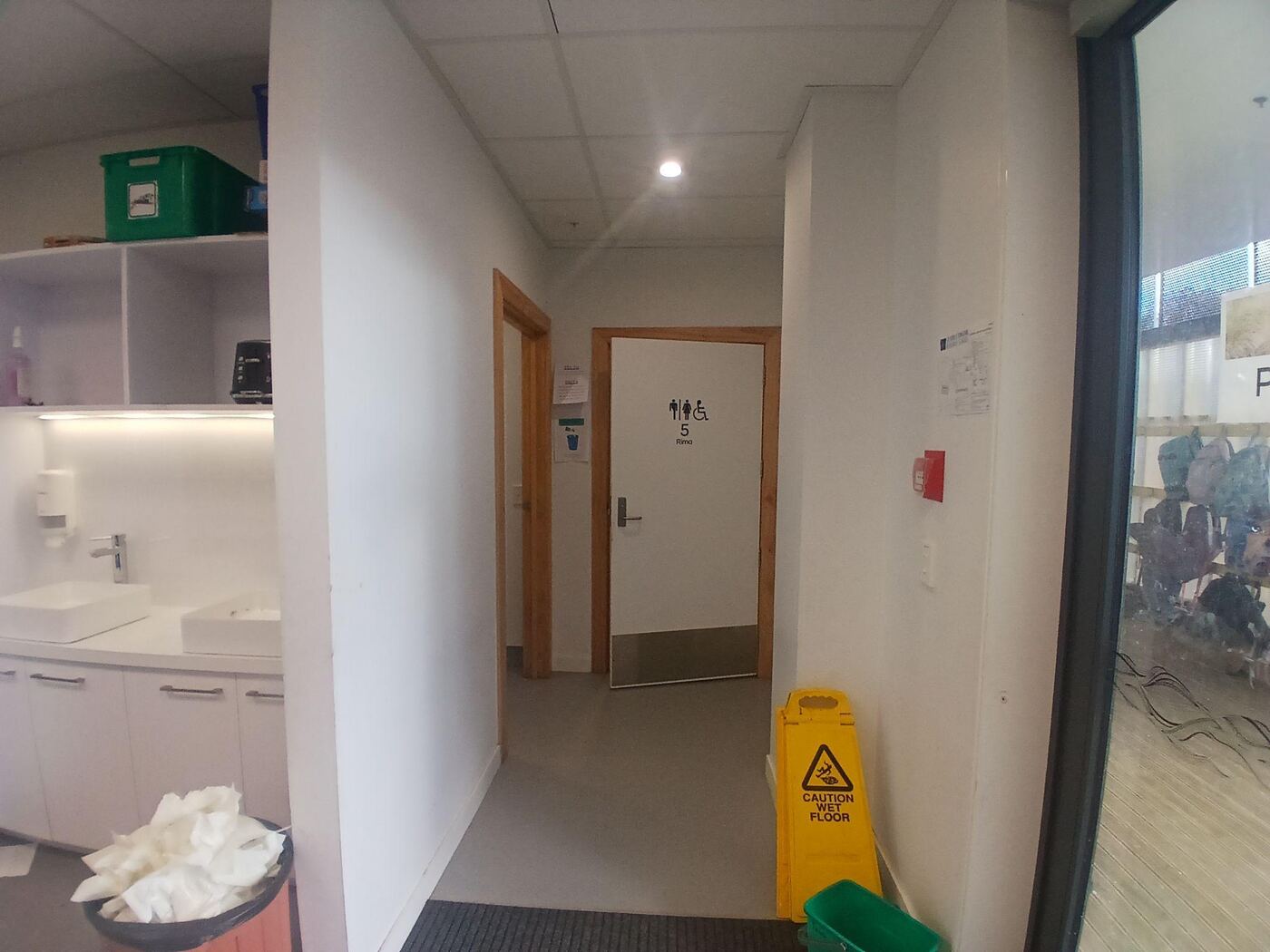 A hallway leading to a door marked with the wheelchair accessible toilet symbol, with wood-toned door frames and a level vinyl floor. A yellow wet-floor sign stands nearby, and a sink and bench are visible in a side room to the left.