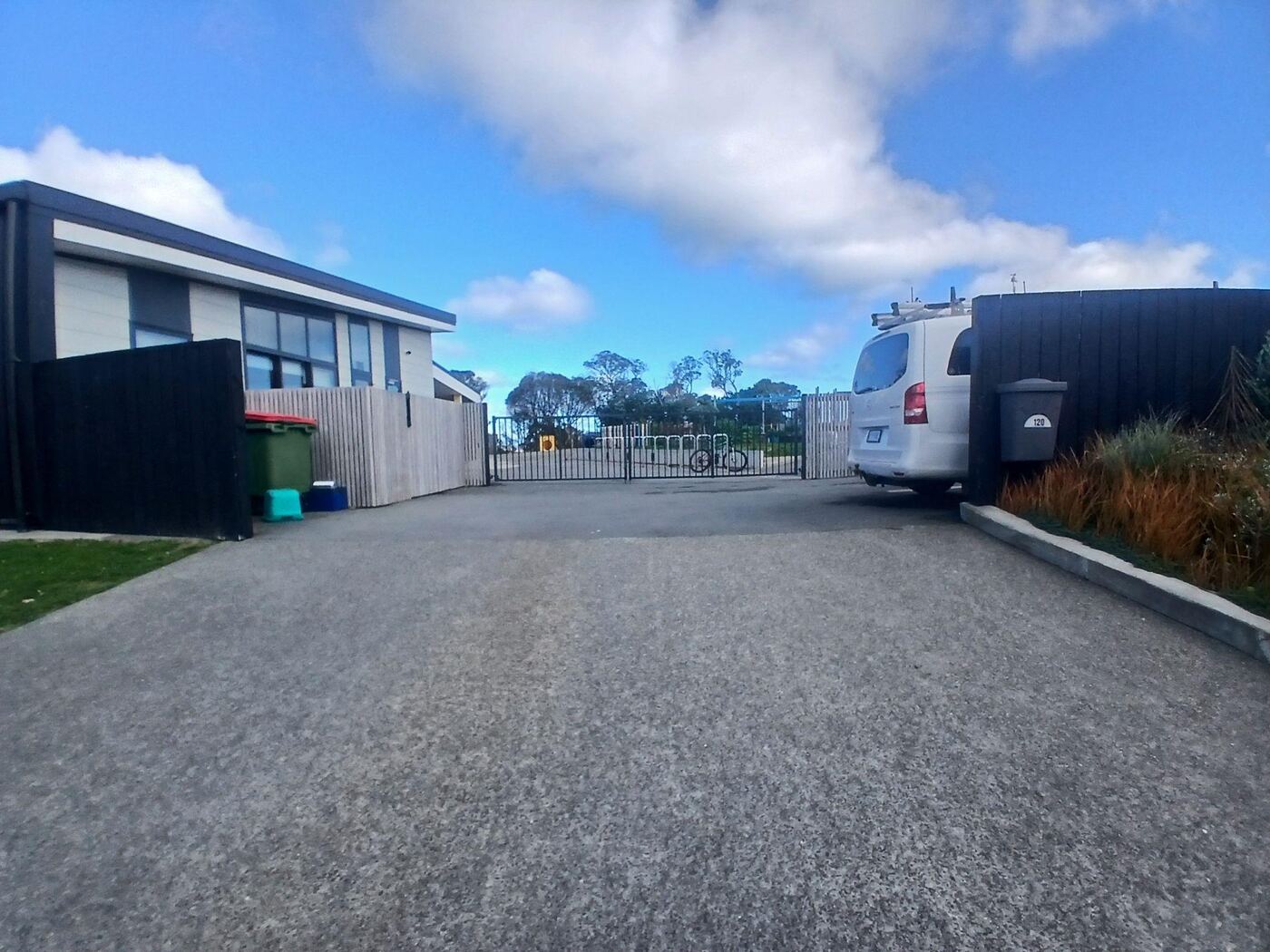 A wide, flat sealed driveway leading up to a black metal gate at Ridgway School, with a van parked to the right and a modern dark-clad school building on the left. A wheelie bin numbered 120 stands beside the gate.