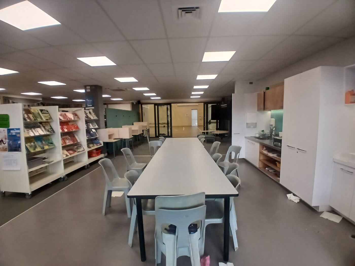 A carpeted room with a long low table and plastic chairs, book display shelves to the left, and a small kitchenette with cupboards and a sink to the right. Glass doors at the far end lead through to another space.