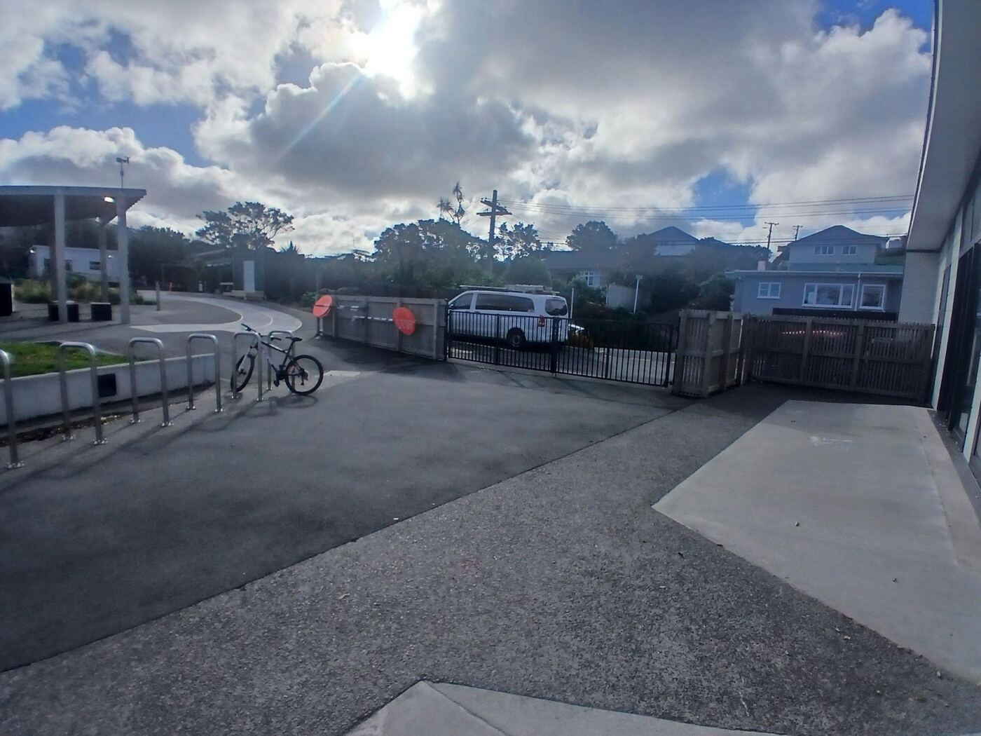 Looking back out through the open school gate towards the road, from inside the grounds. Bike racks and a parked bicycle are to the left, two round orange no-entry signs are fixed to the gate, and the ground is flat sealed asphalt.
