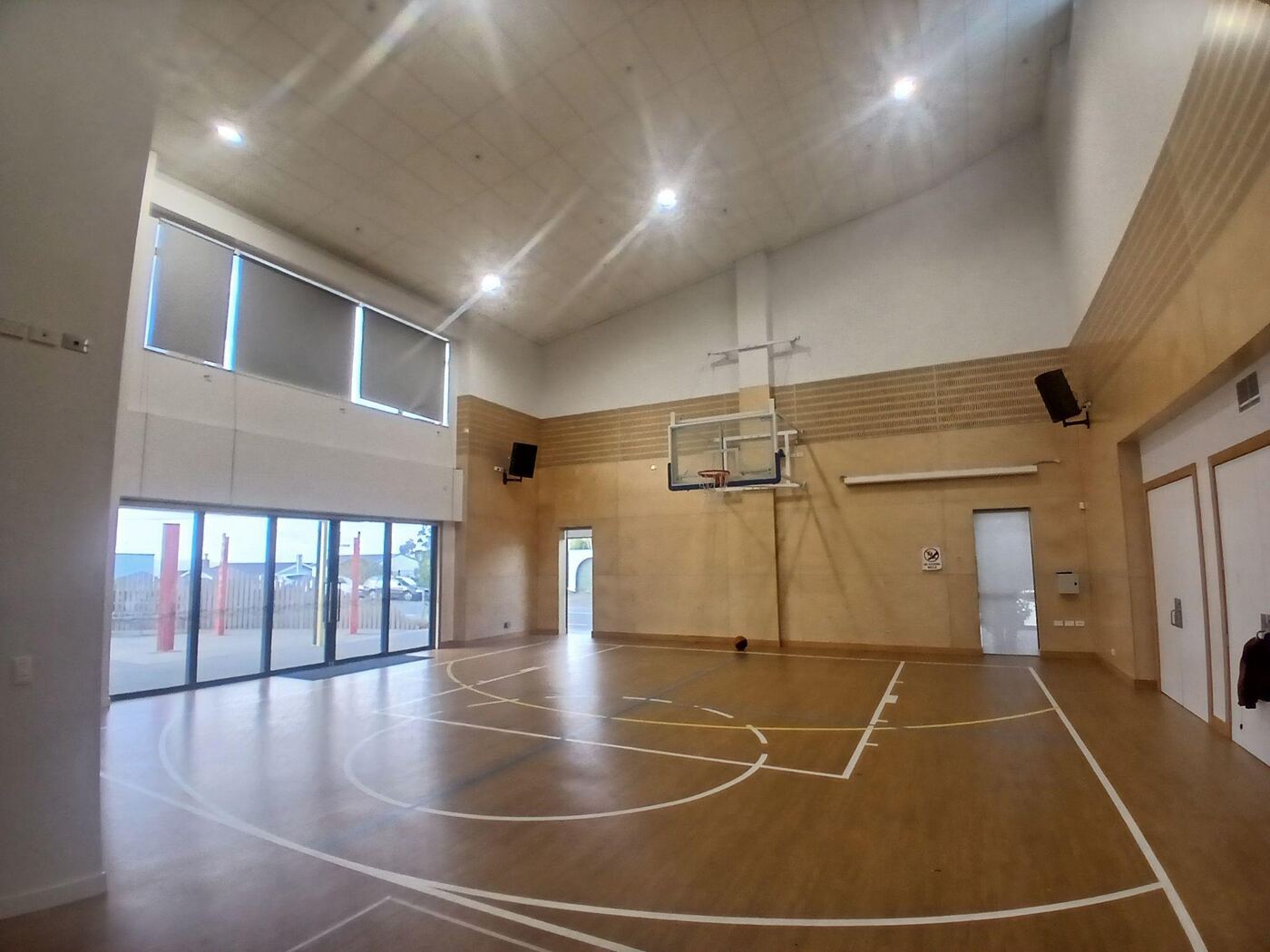 Ridgway School hall: a space like a small gymnasium, with polished wooden floors marked out for basketball, a high ceiling, and floor-to-ceiling glass doors along one side letting in daylight. A basketball hoop is mounted on the far wall.