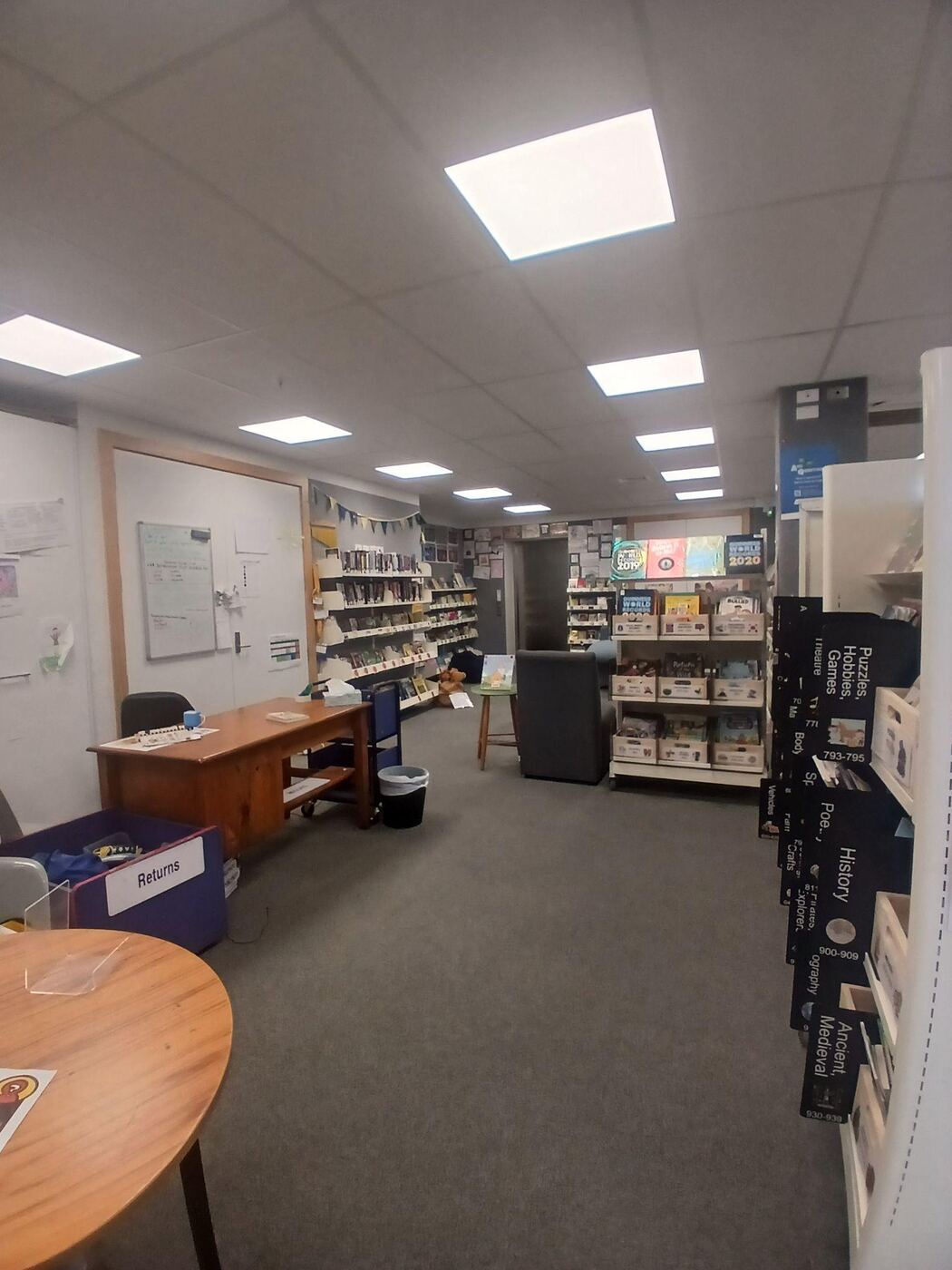 The library nook: grey carpet, a wooden desk, a returns bin, and shelving stocked with books and games along the walls, with a small round table and an armchair in the middle.