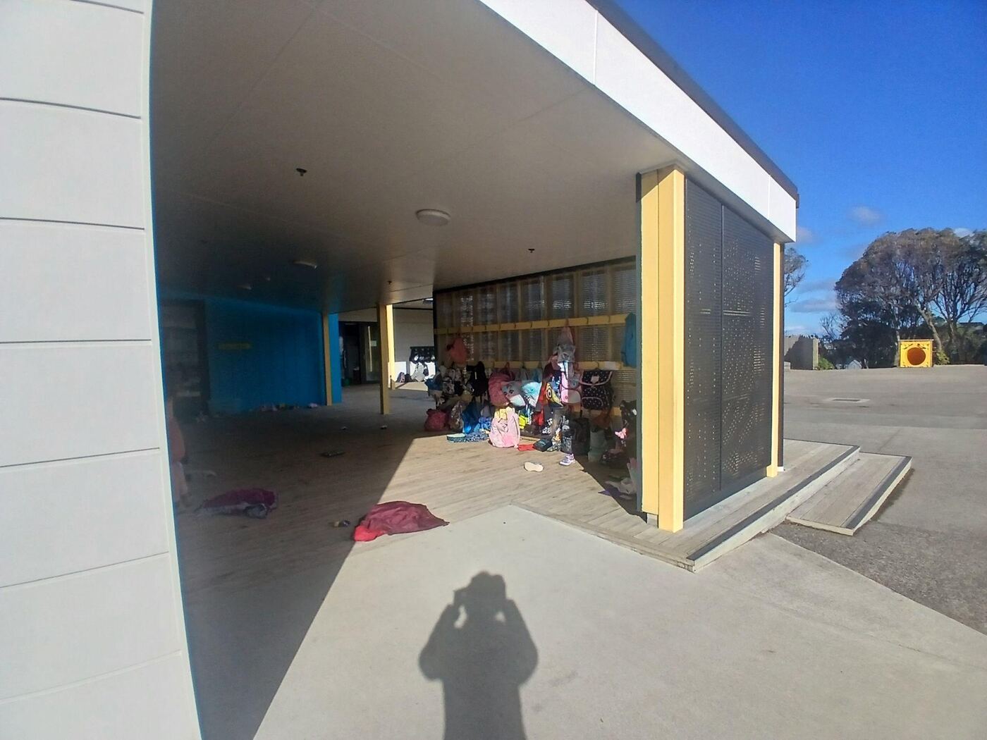 The covered verandah at Ridgway School, with children’s bags and coats hanging along the wall and a wooden deck under the roof. On the right, a concrete ramp with no steps rises gently from the paved courtyard onto the deck.