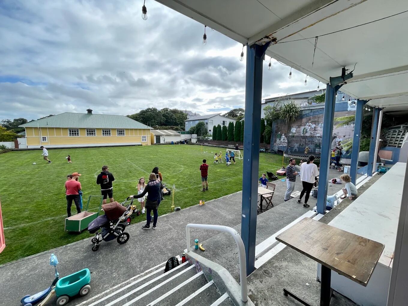 A wide flat grass lawn beside Vogelmorn Bowling Club, seen from a covered deck with blue posts and a step down onto the grass. Folding tables and families with prams are set up along the edge of the lawn, with a soccer goal and the yellow hall building across the field.