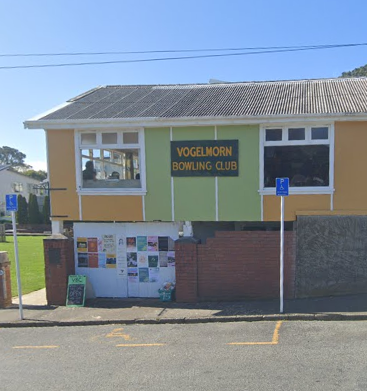 A single car park marked with a wheelchair symbol on the road directly outside Vogelmorn Bowling Club, a yellow and green weatherboard building with a corrugated iron roof. A blue wheelchair-parking sign stands on a pole beside the space, and the club’s gated entrance with noticeboards is a few metres away.