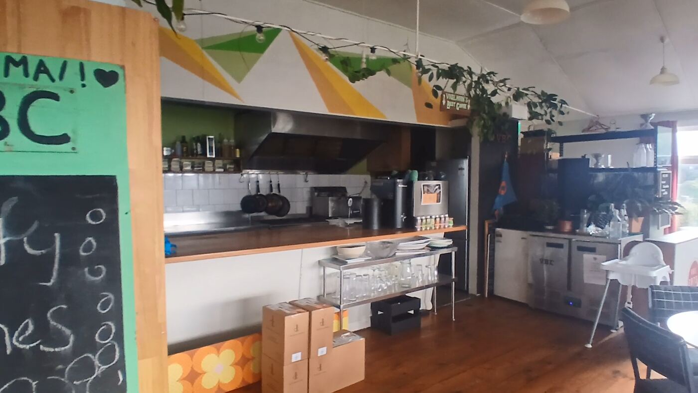 The cafe counter: a long wooden bench with a stainless steel servery, hanging pots and pans, and shelves of glasses and crockery underneath. A high chair stands to the right, below a wall painted with a yellow and green triangle mural.