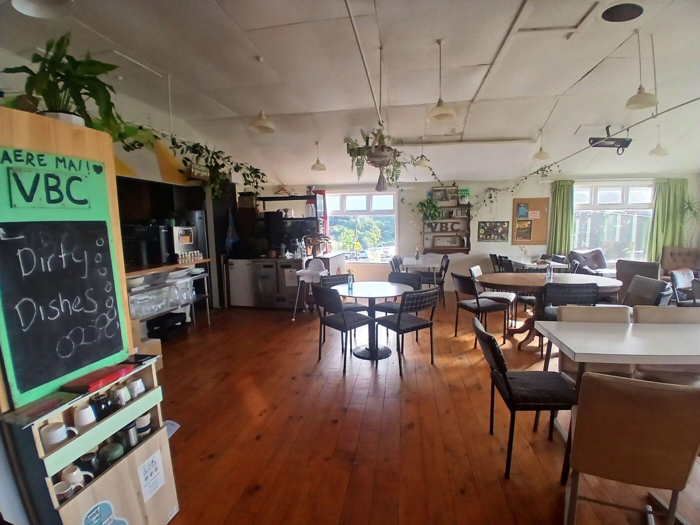The cafe, with polished wooden floorboards, round and square tables with dark upholstered chairs, and hanging pot plants along the ceiling beams. A blackboard sign reading Dirty Dishes stands near a shelf of mugs, with windows and green curtains along the far wall.