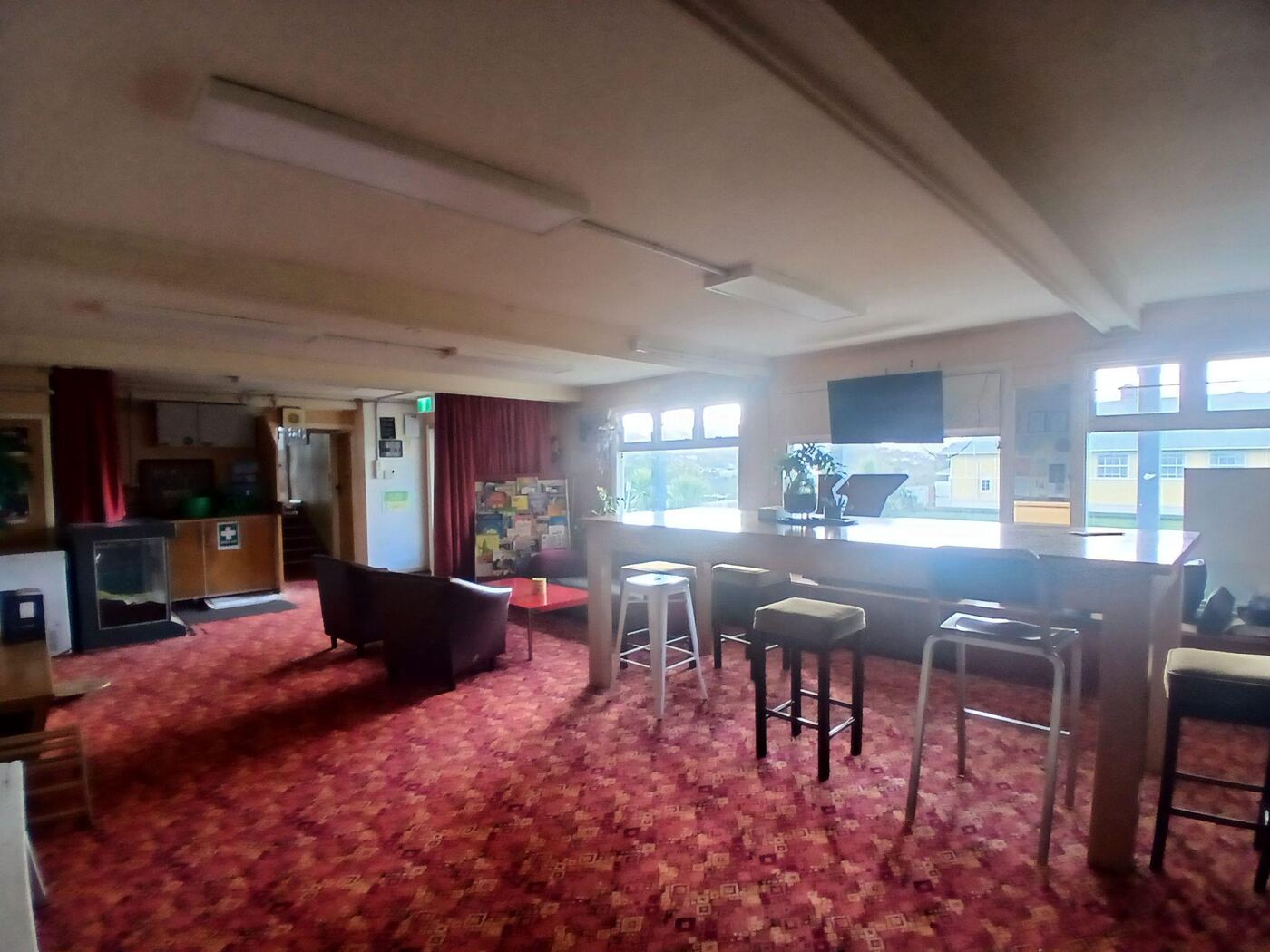 A room with dark red patterned carpet, a black leather couch and a small red coffee table, and a long wooden bar-height table with stools. Large windows along the far wall look out over neighbouring rooftops, and a doorway with stairs leading up is on the left.