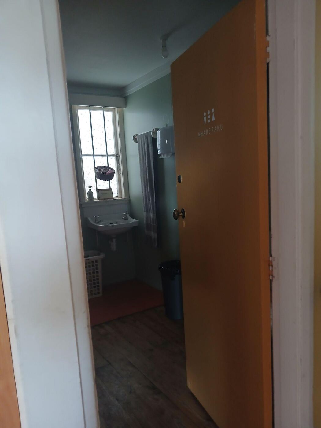 An open wooden door labelled Wharepaku, for all genders, leading into a bathroom with pale green walls, a wall-mounted sink under a window, and a towel rail. The floor is dark wood with a red mat.