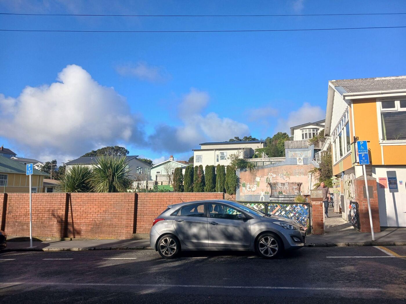 The street outside Vogelmorn Bowling Club, with a car parked at the kerb against a low brick wall. A blue accessible-parking sign stands at one end of the parking area and a 10-minute loading zone sign at the other, beside the gap in the wall that leads to the club’s gate.