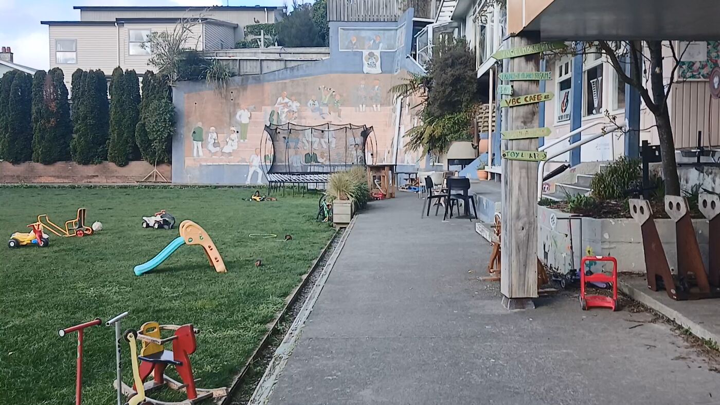 A flat concrete path running alongside the bowling club building. The lawn is on the left, with a trampoline, a slide and ride-on toys on the grass and a painted mural along the far wall. On the right the path passes under a verandah, with wooden steps up to the building and hand-painted signs pointing to the VBC cafe, the toilets and the hall.
