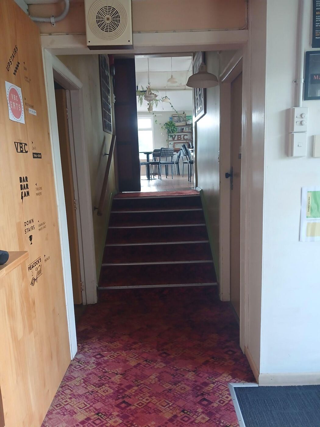 A short flight of six carpeted stairs with a handrail on each side, leading up from a red-carpeted hallway through an open door to cafe seating beyond. A sign on the wall to the left lists the upstairs bar, the downstairs co-working space and the VBC cafe.
