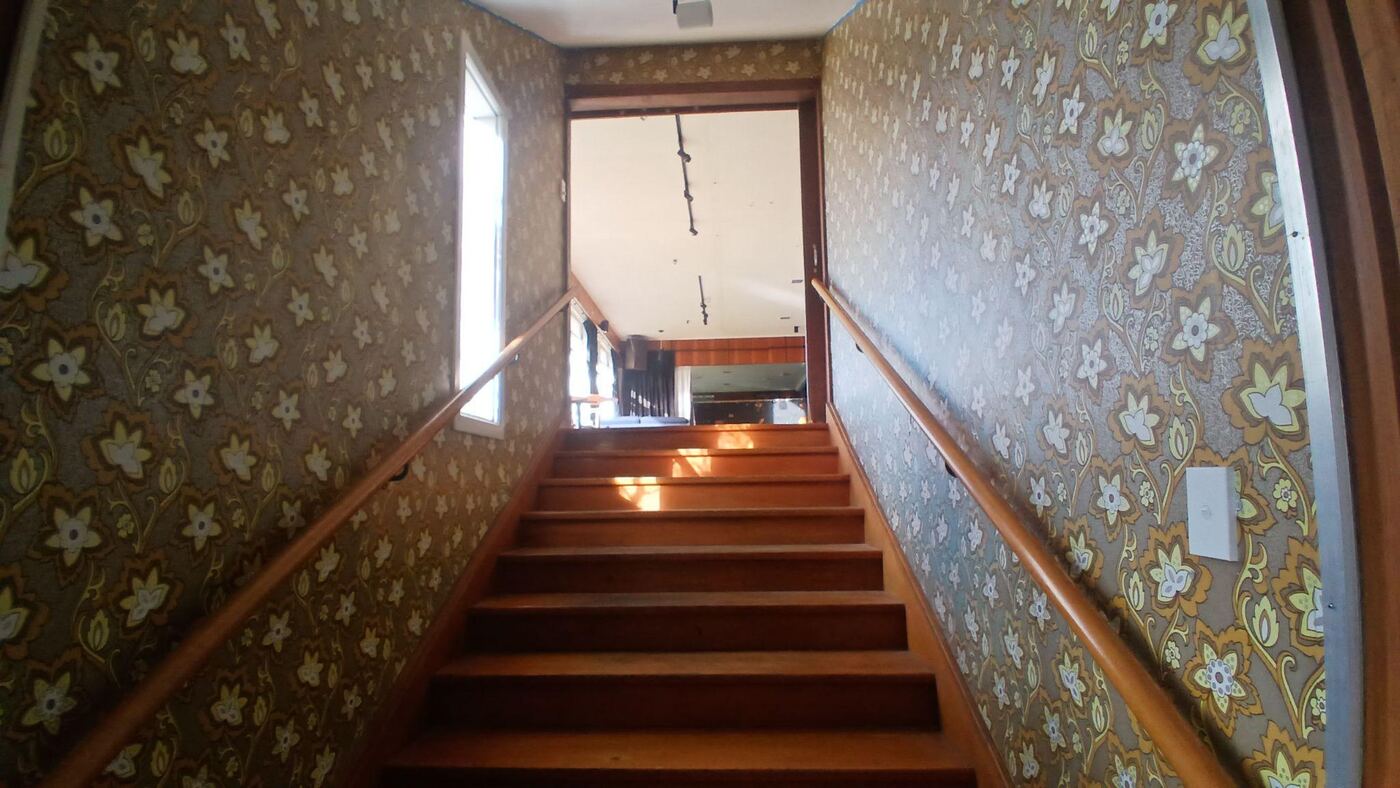A straight flight of about nine wooden stairs with a handrail on each side, between walls covered in dark floral wallpaper, leading up to a bright room with a piano visible through the doorway at the top.