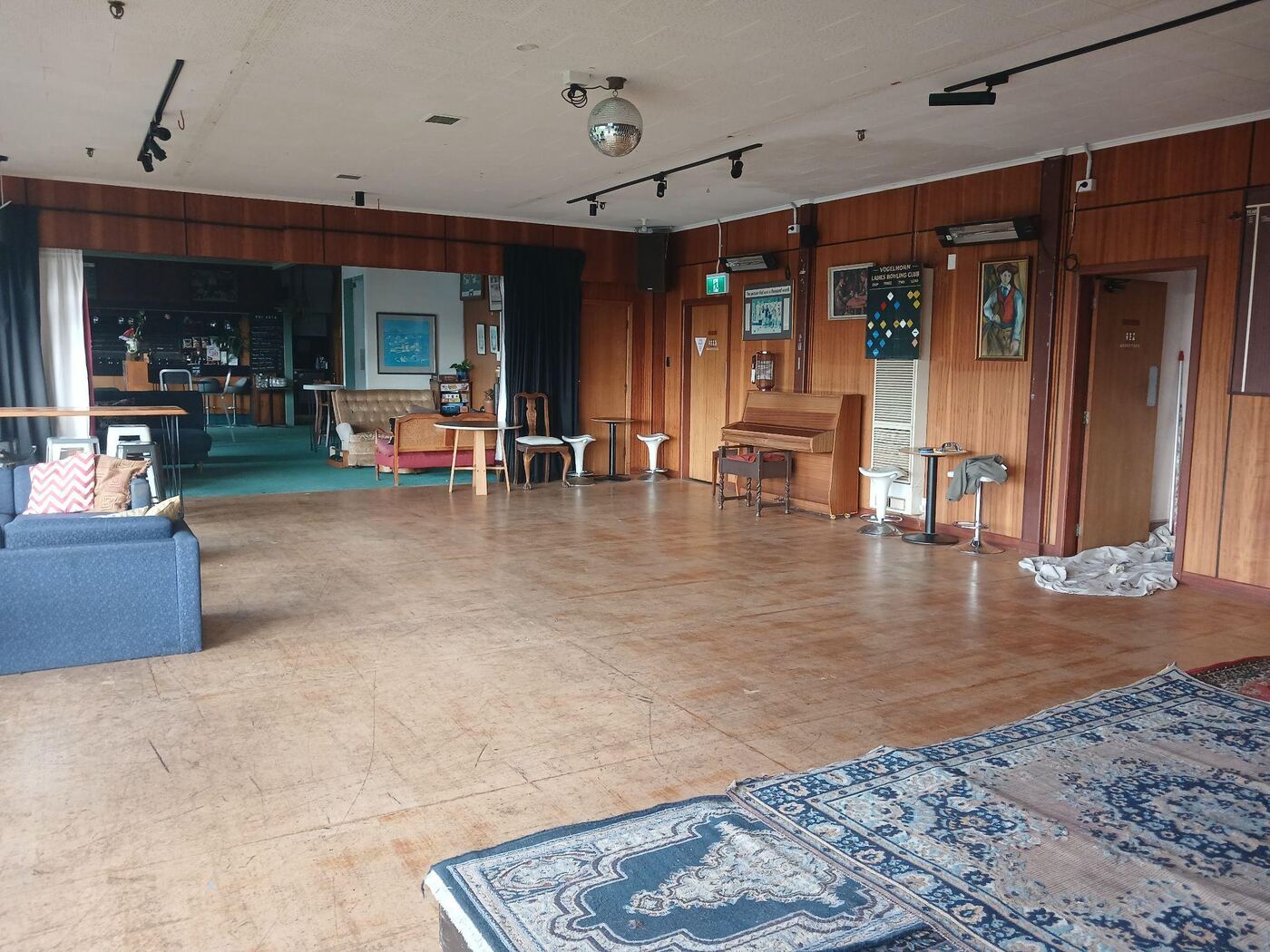 A large room with polished wooden floorboards and dark wood-panelled walls, containing a piano, sofas, bar stools and a disco ball hanging from the ceiling. A dark curtain divides off part of the room, and rugs are laid near the front.